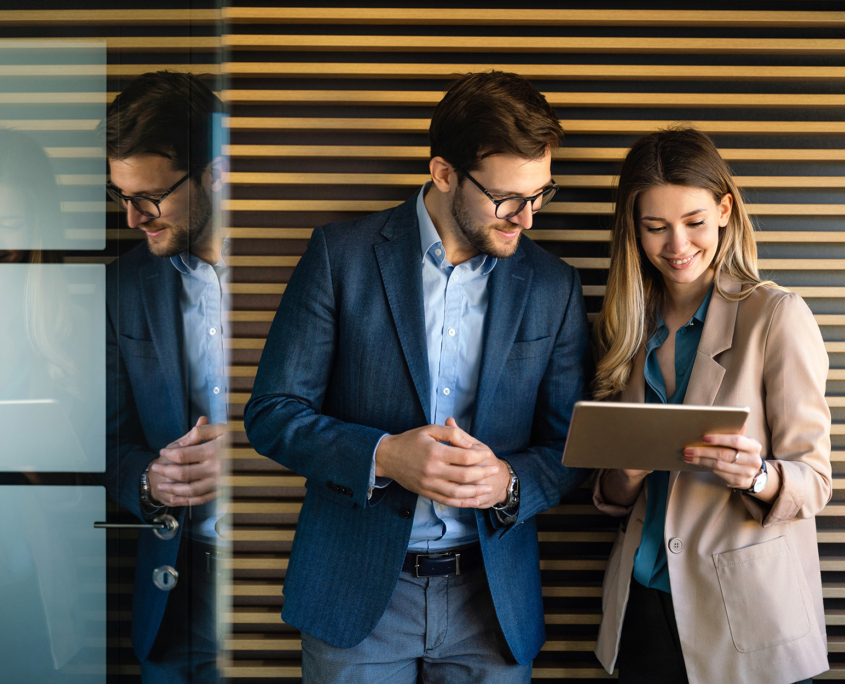 Front View Of Young Male And Female It Professionals Standing In Modern Hallway Looking At Tablet